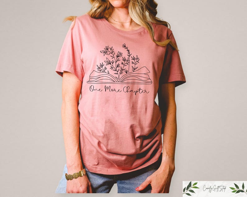 May include: A pink t-shirt with a black line drawing of an open book with flowers growing out of it. The text "One More Chapter" is printed below the book.