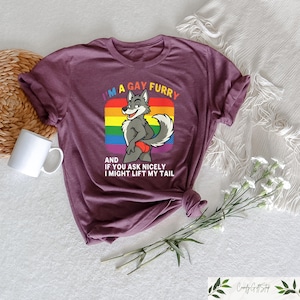 May include: A maroon t-shirt featuring a cartoon wolf character with a rainbow background. The text on the shirt reads "I'M A GAY FURRY AND IF YOU ASK NICELY I MIGHT LIFT MY TAIL". The shirt is folded and has a white mug and flowers nearby.