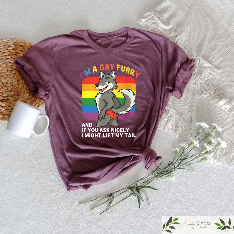 May include: A maroon t-shirt featuring a cartoon wolf character with a rainbow background. The text on the shirt reads "I'M A GAY FURRY AND IF YOU ASK NICELY I MIGHT LIFT MY TAIL". The shirt is folded and has a white mug and flowers nearby.