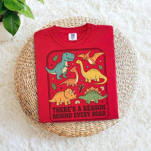 May include: A red t-shirt featuring a colorful dinosaur graphic. The design includes various dinosaurs in shades of green, yellow, and orange, with the text "THERE'S A REASON BEHIND EVERY ROAR". The shirt is folded and placed on a woven surface.