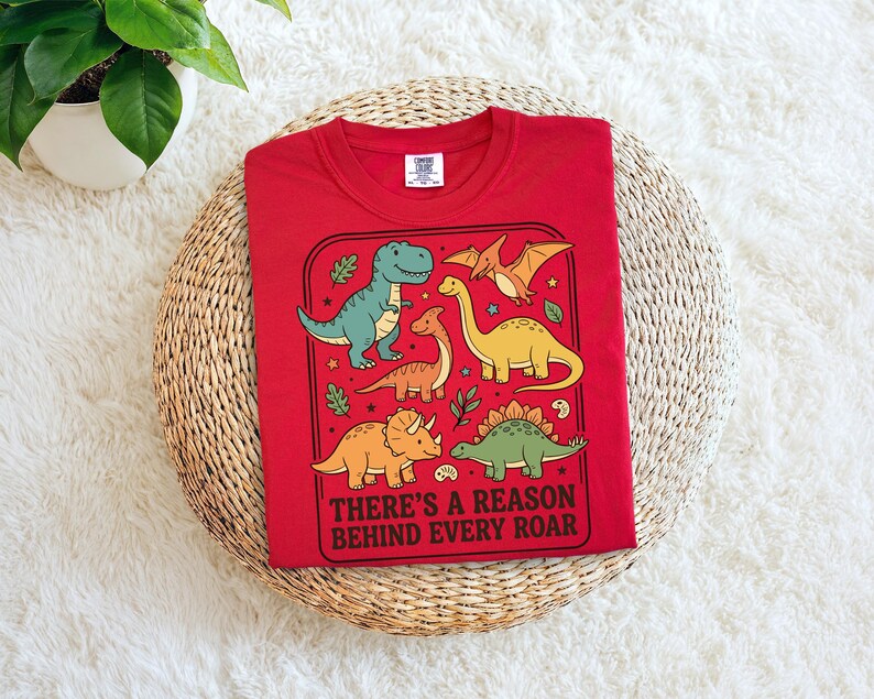 May include: A red t-shirt featuring a colorful dinosaur graphic. The design includes various dinosaurs in shades of green, yellow, and orange, with the text "THERE'S A REASON BEHIND EVERY ROAR". The shirt is folded and placed on a woven surface.