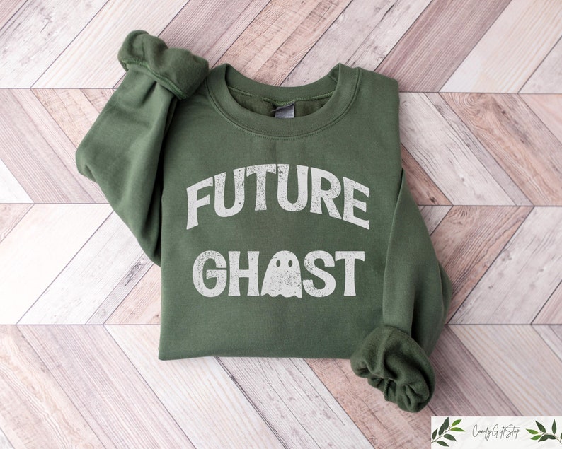 May include: A dark green sweatshirt with the words "FUTURE GHOST" printed in white, along with a ghost graphic. The sweatshirt is folded on a light wood-grain surface.