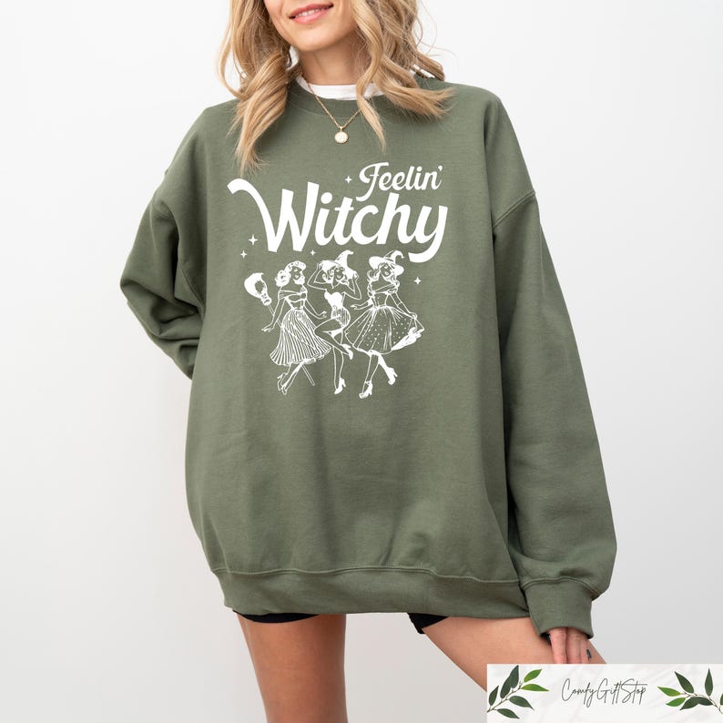 Puede incluir: Sudadera verde oliva con la frase "Feelin' Witchy" en blanco sobre un gr&aacute;fico de tres brujas bailando. La sudadera es de corte holgado y tiene mangas largas.