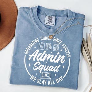 May include: A blue Comfort Colors t-shirt with the text "Organizing Chaos Since Forever Admin Squad We Slay All Day" in white. The shirt has a crew neck and short sleeves. A gold necklace and a brown hat are also in the image.