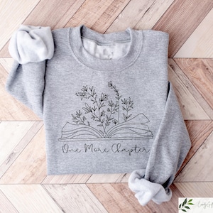 May include: A gray sweatshirt with a black and white illustration of an open book with flowers growing out of it. The text "One More Chapter" is printed below the illustration.