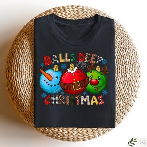 以下が含まれることがあります： クリスマスをテーマにしたグラフィックが特徴の黒いTシャツ。デザインには、雪だるま、サンタ、トナカイの3つのオーナメント型のキャラクターが含まれています。「BALLS DEEP CHRISTMAS」の文字がオーナメントの上と下に表示されています。