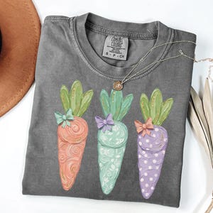 May include: A gray Comfort Colors t-shirt featuring three colorful carrot designs. Each carrot has a different color and pattern, with green tops and bows. A silver necklace with a pendant is draped across the shirt.
