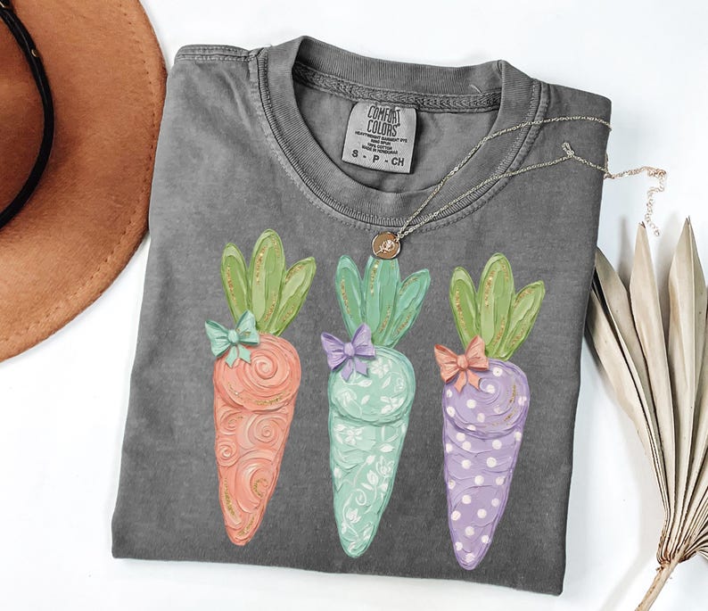 May include: A gray Comfort Colors t-shirt featuring three colorful carrot designs. Each carrot has a different color and pattern, with green tops and bows. A silver necklace with a pendant is draped across the shirt.