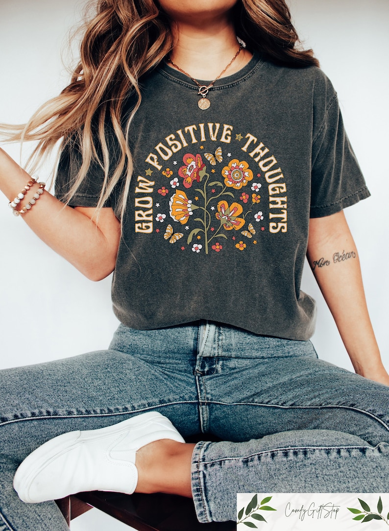 Pu&ograve; includere: Una maglietta grigia scura con un design floreale vintage e il testo "Grow Positive Thoughts".