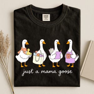 May include: Black t-shirt with a graphic of four cartoon geese, each with a different accessory. The text "just a mama goose" is written in cursive below the geese. The shirt has a Comfort Colors tag.