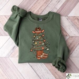 Pu&ograve; includere: Una felpa verde con un disegno di albero di Natale. L'albero indossa un cappello da cowboy e stivali. L'albero &egrave; decorato con ornamenti e luci.