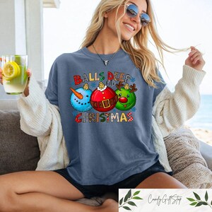 以下が含まれることがあります： クリスマスオーナメントのデザインが施された青いTシャツ。デザインは、雪だるま、サンタクロース、トナカイの3つのオーナメントが特徴です。「BALLS DEEP in CHRISTMAS」の文字がオーナメントの上と下にプリントされています。