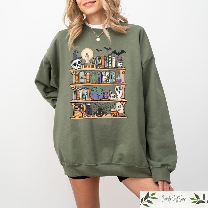 May include: A green sweatshirt featuring a graphic of a bookshelf filled with books, potions, and Halloween decorations. The bookshelf is decorated with bats, a skull, a cauldron, and a ghost.