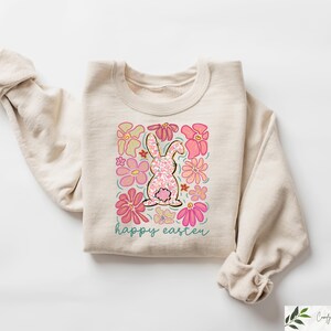 Peut inclure: Un sweat-shirt beige avec un lapin rose entour&eacute; de fleurs roses. Le texte "happy easter" est imprim&eacute; sous le lapin.