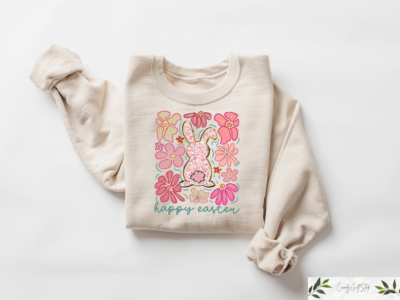 Peut inclure: Un sweat-shirt beige avec un lapin rose entour&eacute; de fleurs roses. Le texte "happy easter" est imprim&eacute; sous le lapin.