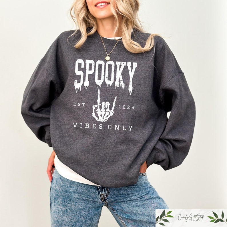 Puede incluir: Una sudadera gris oscura con un estampado gr&aacute;fico blanco que dice "Spooky Est. 1629 Vibes Only". El gr&aacute;fico presenta una mano de esqueleto haciendo el dedo medio.