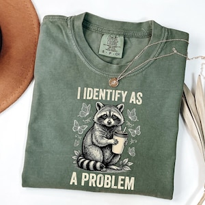 May include: An olive green crew neck t-shirt featuring a graphic of a raccoon holding a coffee cup surrounded by butterflies. The text "I IDENTIFY AS A PROBLEM" is printed in a cream-coloured font above and below the raccoon illustration.