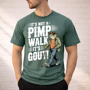 May include: Green t-shirt with the text "IT'S NOT A PIMP WALK IT'S GOUT!" and a cartoon frog wearing sunglasses, a plaid shirt, jeans, and sneakers, walking with a cane. The frog is the central graphic.