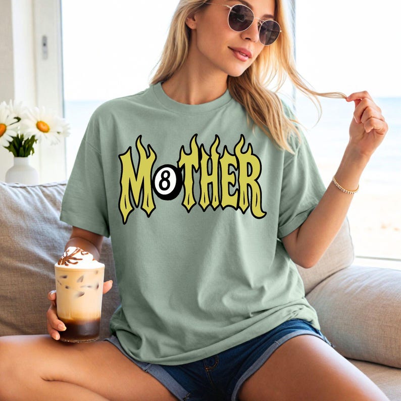 May include: Sage green t-shirt with the word "MOTHER" in yellow, flame-like lettering, and an 8-ball graphic. The shirt is a relaxed fit and has short sleeves. The model is wearing blue jean shorts.