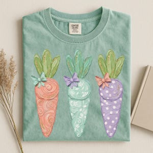 May include: A light green t-shirt featuring three decorative carrots. Each carrot is a different color: peach, mint, and lavender, with green tops and bows. The peach carrot has gold swirls, the mint carrot has a floral pattern, and the lavender carrot has white polka dots.