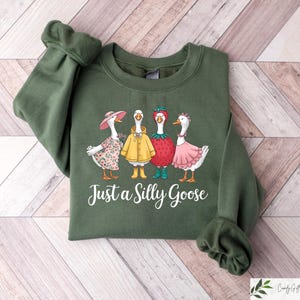 May include: A green sweatshirt with a graphic of four geese wearing different outfits. The geese are wearing a flower crown, a yellow raincoat, a strawberry hat, and a pink dress. The text "Just a Silly Goose" is printed below the geese.