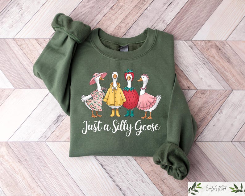 May include: A green sweatshirt with a graphic of four geese wearing different outfits. The geese are wearing a flower crown, a yellow raincoat, a strawberry hat, and a pink dress. The text "Just a Silly Goose" is printed below the geese.