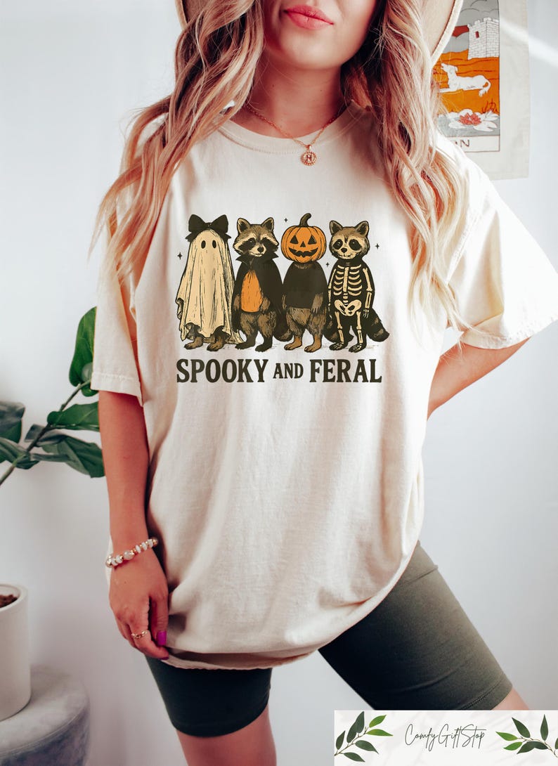 May include: A cream-coloured t-shirt with a Halloween graphic. The design includes a ghost, two raccoons, a pumpkin-headed figure, and a skeleton raccoon. The text "SPOOKY AND FERAL" is printed beneath the characters.