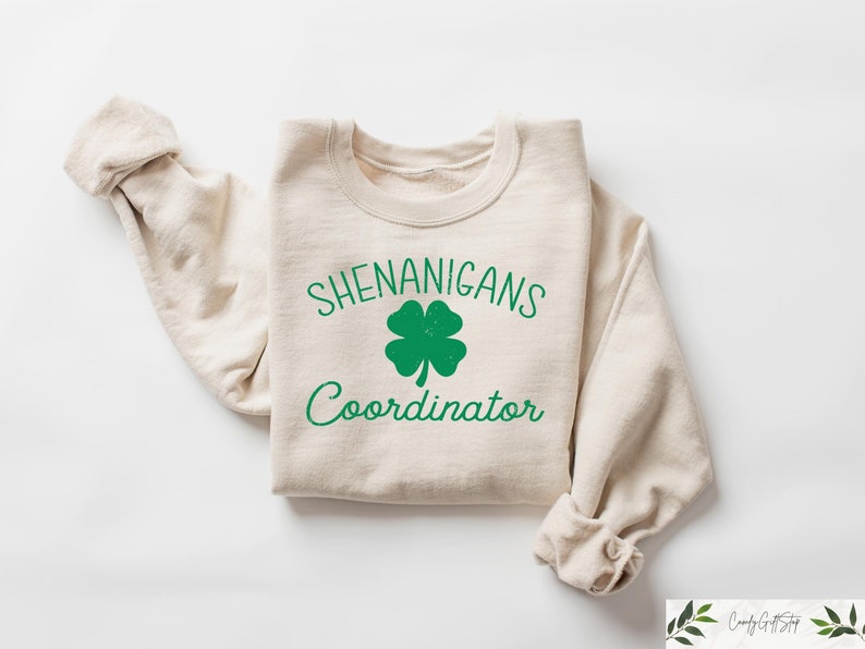 May include: A beige sweatshirt with the text "Shenanigans Coordinator" and a green four-leaf clover graphic.