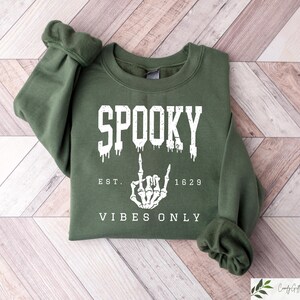 Puede incluir: Una sudadera verde con el texto "Spooky Est. 1629 Vibes Only" y una mano de esqueleto blanca haciendo un gesto de rock and roll.