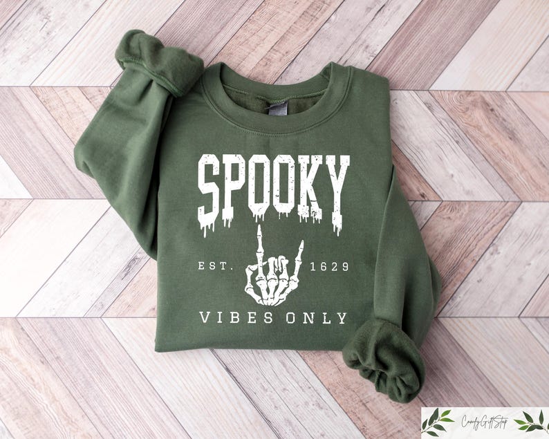 Puede incluir: Una sudadera verde con el texto "Spooky Est. 1629 Vibes Only" y una mano de esqueleto blanca haciendo un gesto de rock and roll.