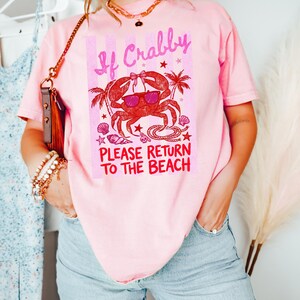 May include: Pink t-shirt with a crab graphic wearing sunglasses and the text "If Crabby Please Return to the Beach." The crab design is red with palm trees and seashells. The shirt is a casual, beach-themed apparel.