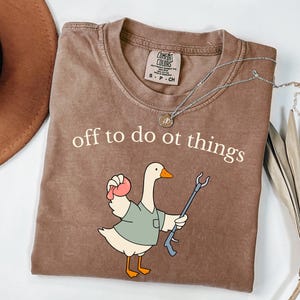 May include: A brown short-sleeve t-shirt featuring a graphic of a goose wearing a green shirt, holding a pink object and a reacher grabber tool. The text above the goose reads, off to do ot things. The shirt has a Comfort Colors brand label.
