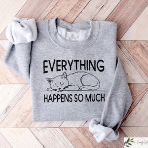 K&ouml;nnte beinhalten: Ein graues Sweatshirt mit dem Text "EVERYTHING HAPPENS SO MUCH" &uuml;ber einer Illustration einer schlafenden Katze. Das Sweatshirt ist auf einer hellbraunen Holzoberfl&auml;che gefaltet.