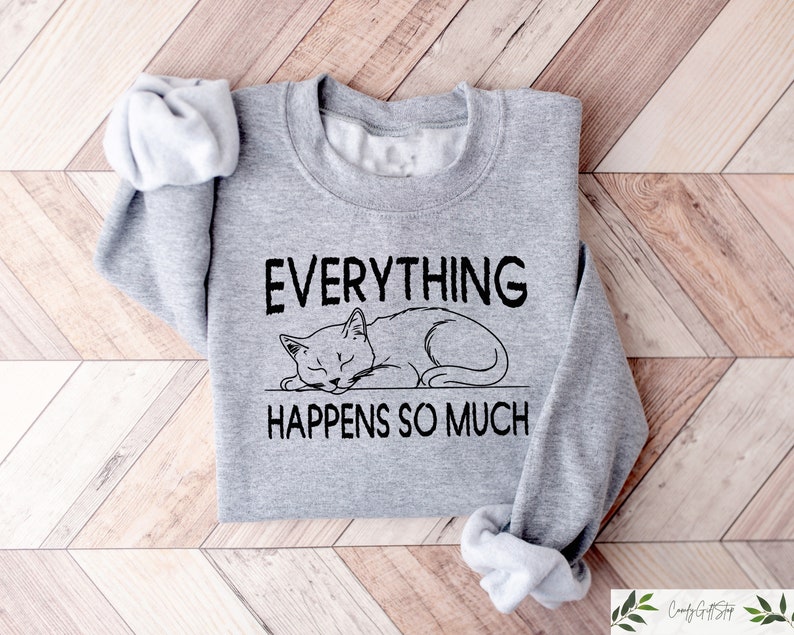 K&ouml;nnte beinhalten: Ein graues Sweatshirt mit dem Text "EVERYTHING HAPPENS SO MUCH" &uuml;ber einer Illustration einer schlafenden Katze. Das Sweatshirt ist auf einer hellbraunen Holzoberfl&auml;che gefaltet.