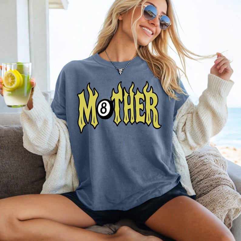 May include: A blue t-shirt with the word "MOTHER" in yellow flame-like lettering and the number 8 ball graphic. The shirt is worn by a person sitting outdoors.