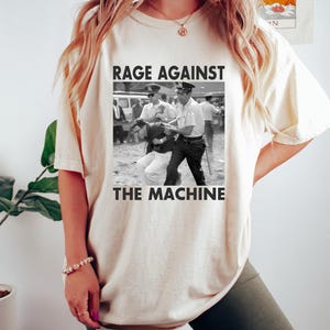 May include: Cream-colored t-shirt featuring a black and white image of a protest scene with the text "RAGE AGAINST THE MACHINE". The shirt has short sleeves and a relaxed fit, suitable for casual wear.