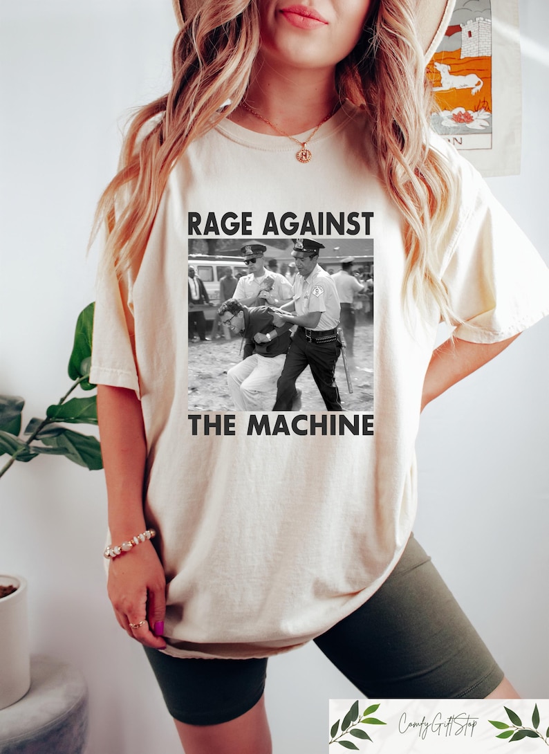 May include: Cream-colored t-shirt featuring a black and white image of a protest scene with the text "RAGE AGAINST THE MACHINE". The shirt has short sleeves and a relaxed fit, suitable for casual wear.