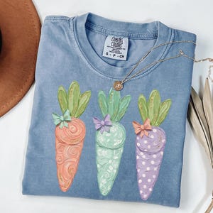 May include: A blue Comfort Colors t-shirt featuring three colorful carrot designs. Each carrot has a different pattern and bow. The carrots are orange, mint green, and purple. A gold necklace with a small pendant is draped across the shirt.