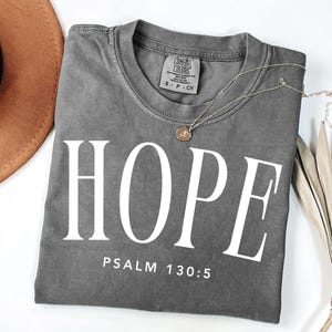Puede incluir: Una camiseta gris Comfort Colors con la palabra "HOPE" impresa en letras blancas grandes. Debajo de la palabra "HOPE" está el texto "PSALM 130:5". Un collar dorado con un colgante está drapeado sobre la camisa. Un sombrero marrón y una planta seca están en el fondo.