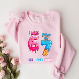 May include: A light pink sweatshirt with the text "I WAS 67 NOW I'M 7 SIX SEVEN BRUH" in colorful, cartoon-style lettering. The number 6 is designed as a donut, and the number 7 as an ice cream cone.