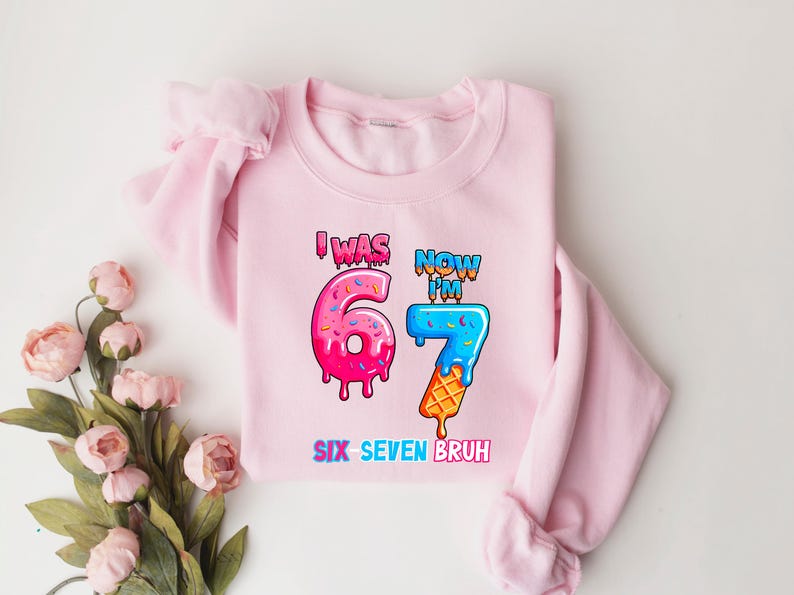 May include: A light pink sweatshirt with the text "I WAS 67 NOW I'M 7 SIX SEVEN BRUH" in colorful, cartoon-style lettering. The number 6 is designed as a donut, and the number 7 as an ice cream cone.