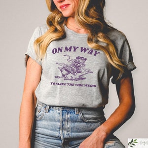 May include: A gray t-shirt with a purple graphic of a person riding a bird. The text on the shirt reads "On My Way To Make The Vibe Weird."
