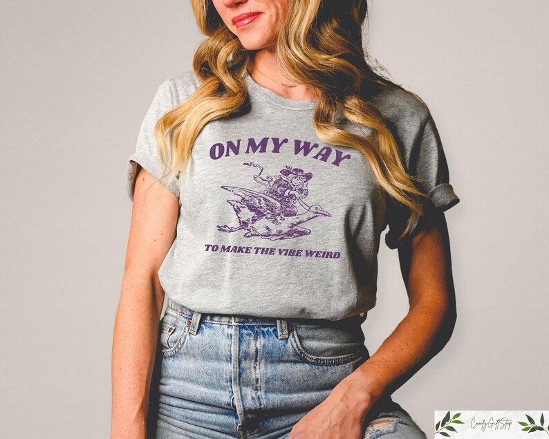 May include: A gray t-shirt with a purple graphic of a person riding a bird. The text on the shirt reads "On My Way To Make The Vibe Weird."