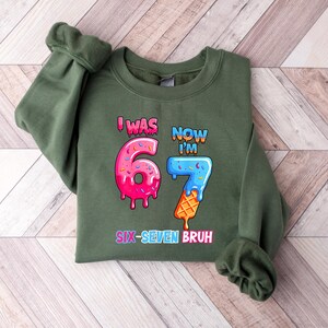 May include: An olive green sweatshirt with the text "I WAS 6, NOW I'M 7" in colorful, cartoonish lettering. The number 6 is designed as a pink donut, and the number 7 as a blue ice cream cone. The phrase "SIX-SEVEN BRUH" is at the bottom.
