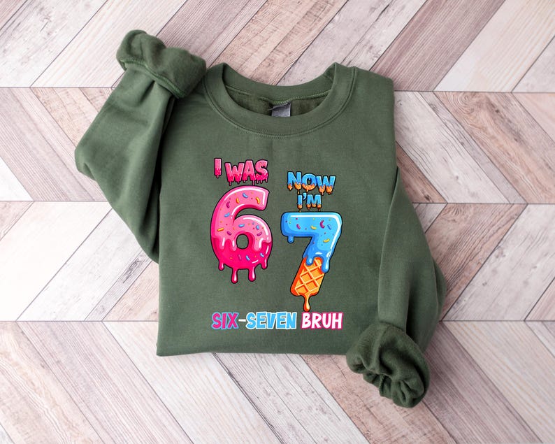 May include: An olive green sweatshirt with the text "I WAS 6, NOW I'M 7" in colorful, cartoonish lettering. The number 6 is designed as a pink donut, and the number 7 as a blue ice cream cone. The phrase "SIX-SEVEN BRUH" is at the bottom.