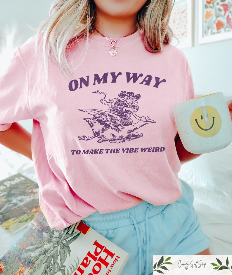 May include: Pink t-shirt with a purple graphic of a person riding a goose. The text on the shirt reads "On my way to make the vibe weird."