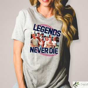 May include: A light gray t-shirt featuring a graphic from the movie "The Sandlot." The graphic includes a baseball with the text "Legends Never Die" and a photo of the movie's cast. The shirt is a crew neck style.