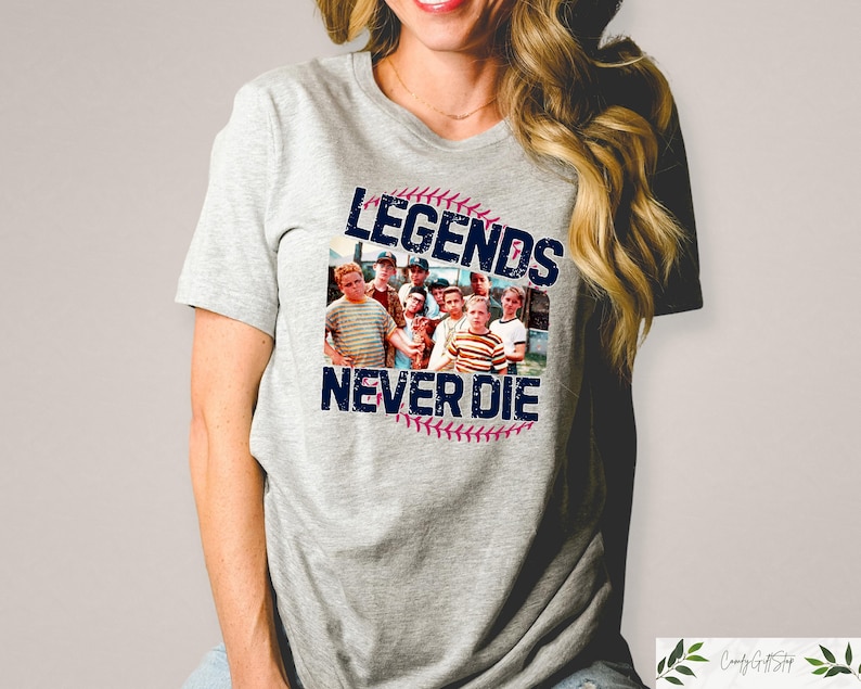 May include: A light gray t-shirt featuring a graphic from the movie "The Sandlot." The graphic includes a baseball with the text "Legends Never Die" and a photo of the movie's cast. The shirt is a crew neck style.