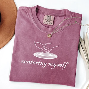 May include: A dusty rose Comfort Colors t-shirt with the words "centering myself" in white script. The shirt features a graphic of hands working with clay on a pottery wheel. A gold necklace with a pendant rests on the shirt.