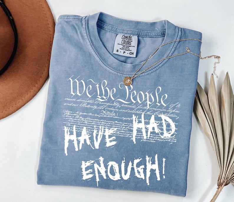 Puede incluir: Una camiseta azul Comfort Colors con el texto "We the People HAVE HAD ENOUGH!" en blanco. Un collar dorado con un colgante descansa sobre la camiseta. Un sombrero marr&oacute;n y hojas de palma secas est&aacute;n en el fondo.